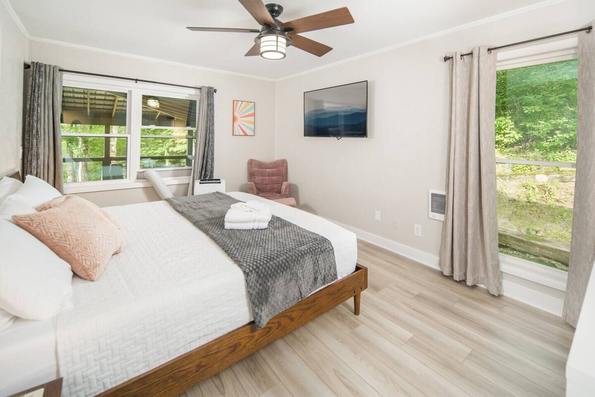 A bright bedroom features a spacious bed with soft linens and neutral decor. A ceiling fan is present, and natural light floods in from large windows. An armchair sits beside the bed, and a wall-mounted TV adds to the room's functionality.
