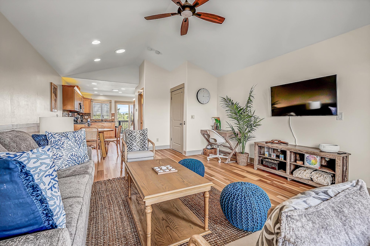 The spacious living area features a light-colored couch and a wooden coffee table, with blue accent pillows and poufs. Natural wood flooring complements the room, while a clock and large TV add modern touches. Light floods in through the open space connecting to the kitchen.