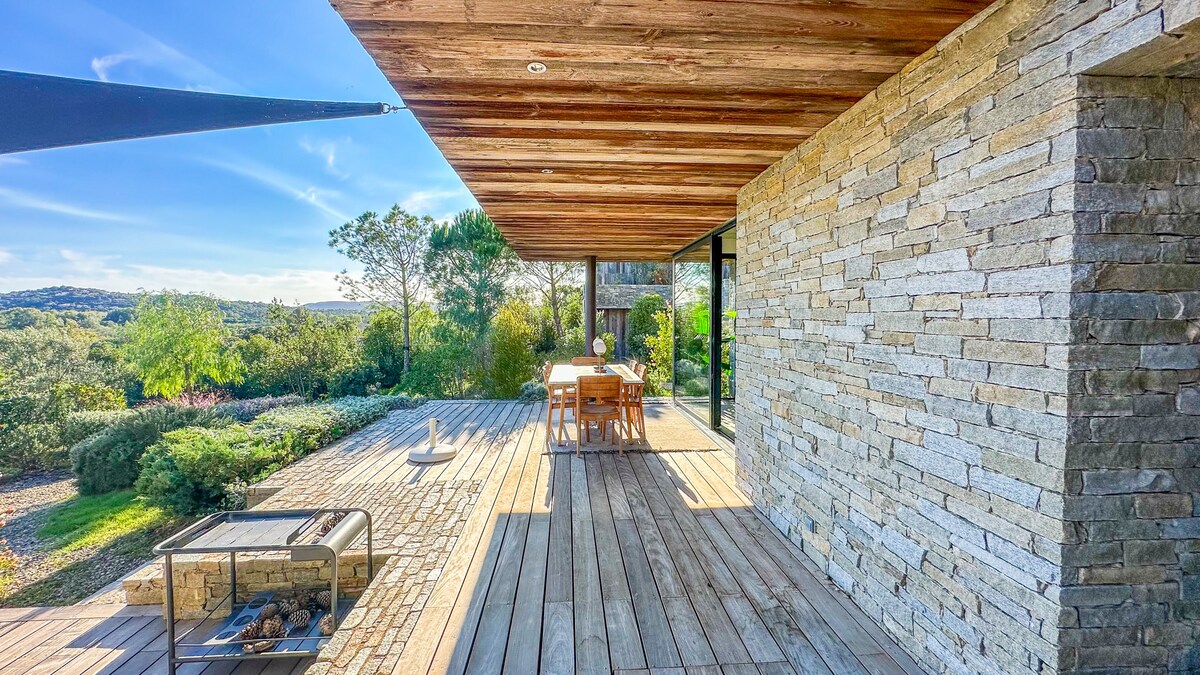 An outdoor terrace features wooden flooring with a stone wall backdrop and a large dining table surrounded by chairs. A shaded area is provided by an extended roof, and a fire pit is visible to the side, surrounded by natural greenery.