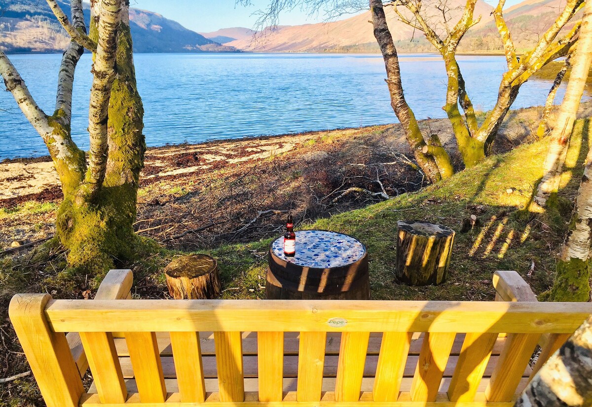 A wooden bench is positioned near the water's edge, offering a view of the calm loch. A small round table is visible, set atop a stone base. The scene is framed by trees with moss-covered trunks, reflecting natural surroundings.