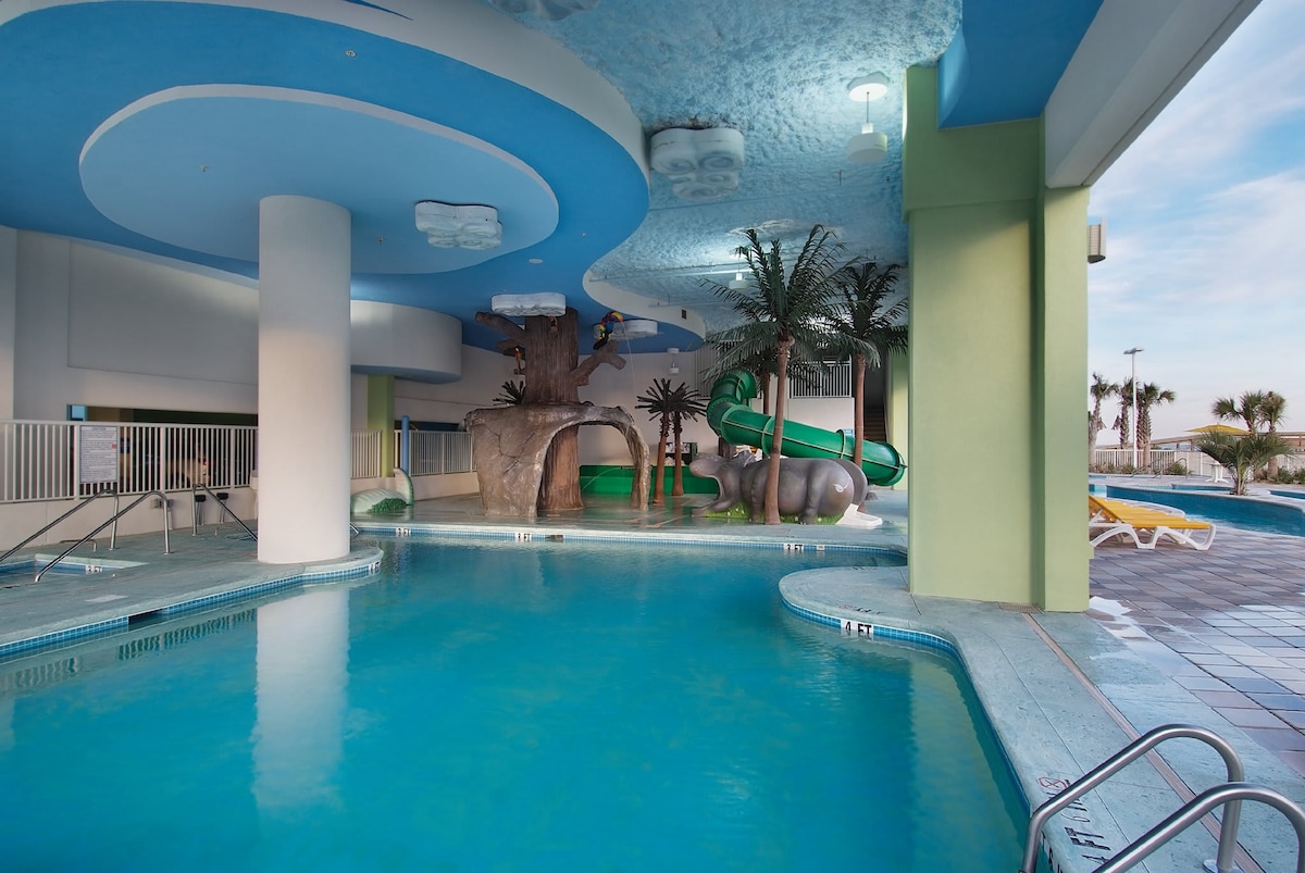 An indoor pool area features a light blue ceiling that mimics water, with playful palm trees and animal sculptures surrounding the space. Water slides can be seen at one edge, and lounge chairs are arranged for relaxation beside the pool.