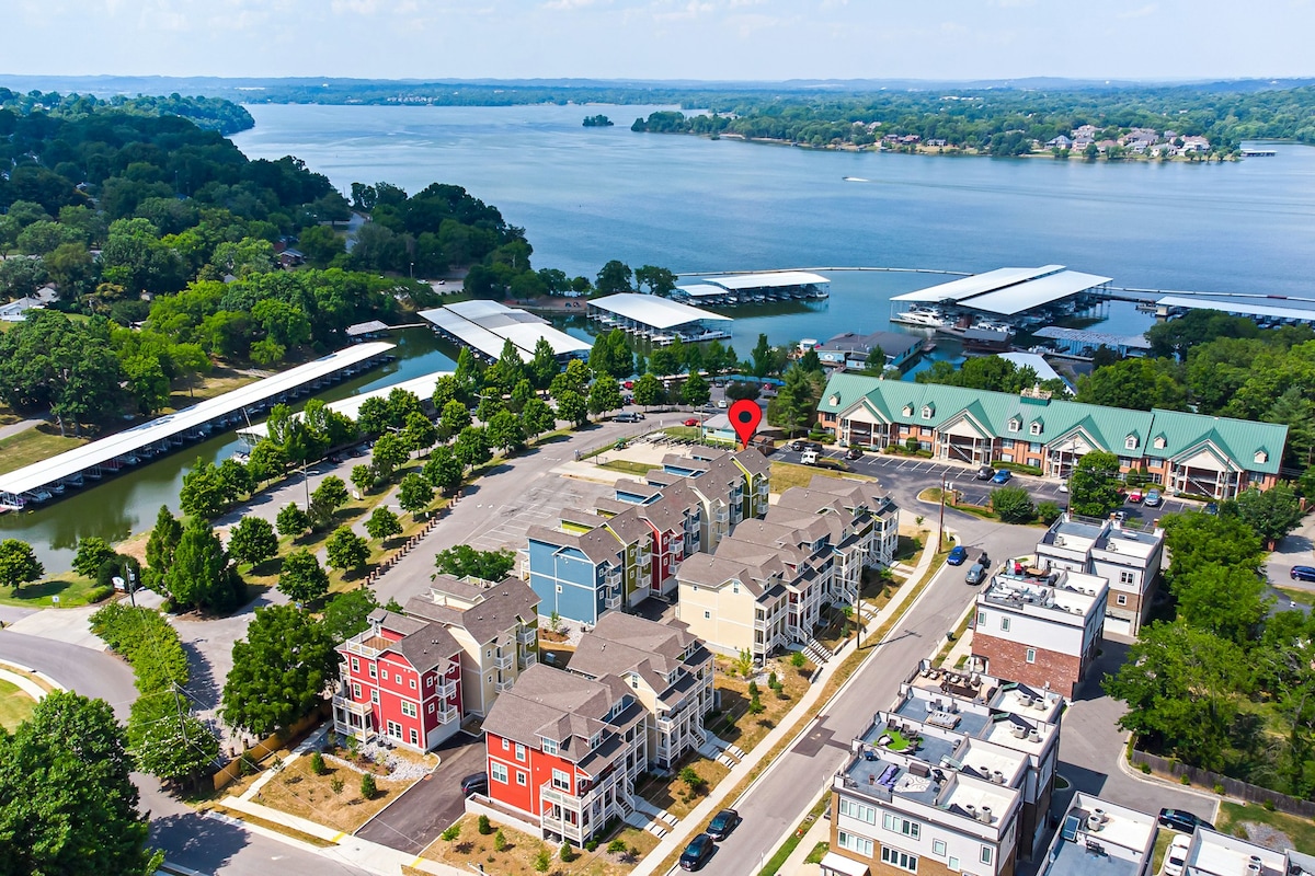An aerial view captures the charming townhome community alongside Old Hickory Lake, featuring several colorful buildings and a marina. The expansive water stretches into the distance, edged by greenery, while the organized parking area and surrounding streets indicate easy access to the townhomes.