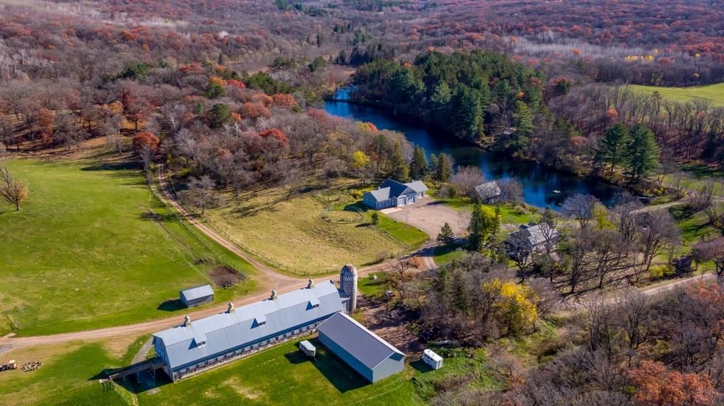 An aerial view captures the expansive property with a large barn, several buildings, and a serene pond surrounded by lush greenery. Autumn foliage adds vibrant colors to the landscape, while gravel paths weave through the open terrain, providing a sense of tranquility.