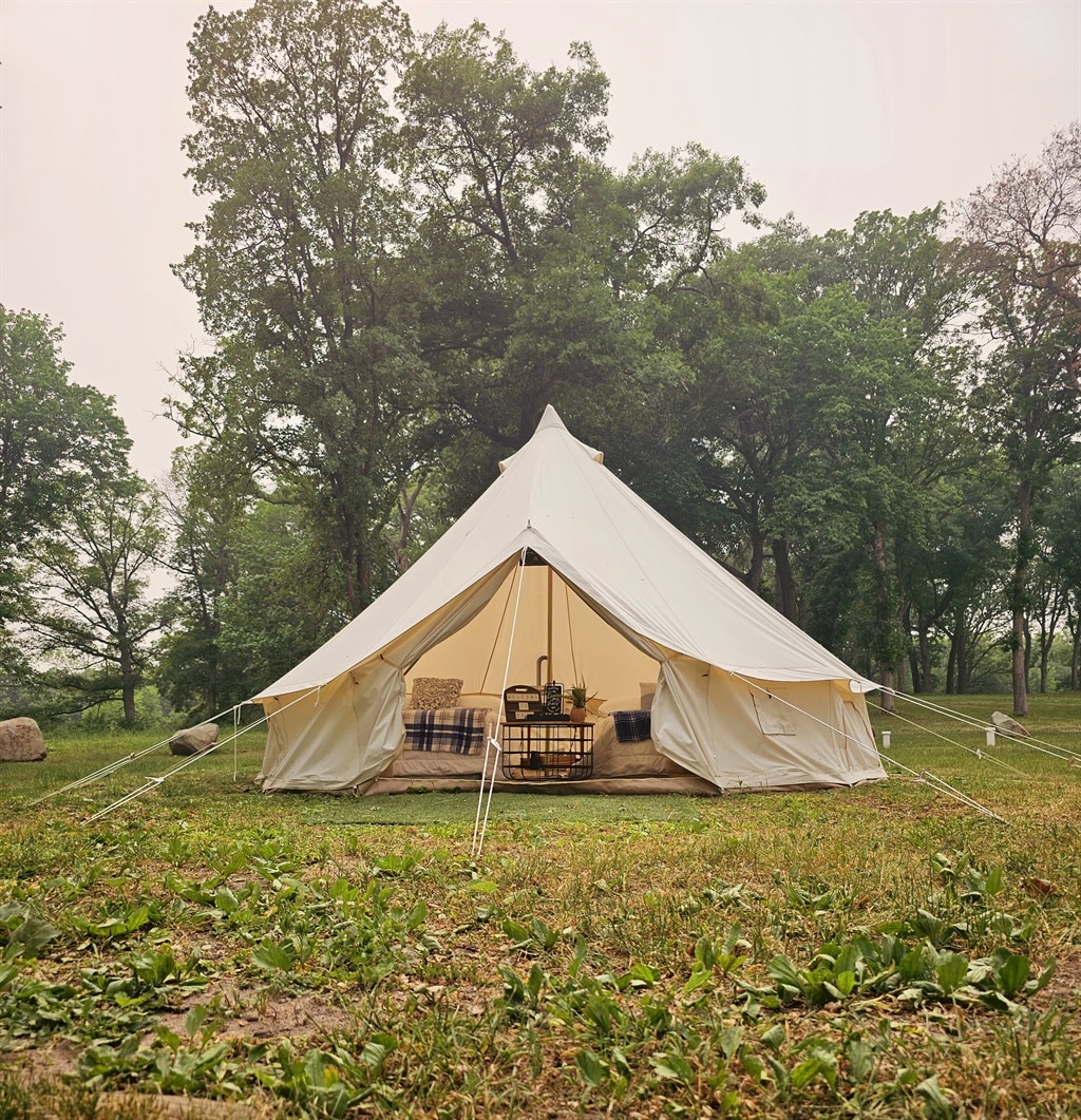 A glamp tent is set among greenery, featuring a white exterior and a spacious entrance. Inside, comfortable seating and a small table are visible, surrounded by nature. Tall trees provide a serene backdrop, emphasizing a tranquil outdoor setting.