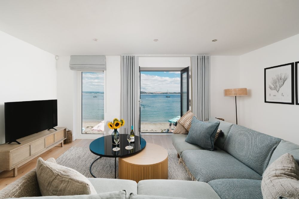 A spacious living area features a corner sofa and an armchair, arranged around a circular coffee table. Large windows and a Juliette balcony provide an unobstructed view of the sea, with natural light enhancing the calming color palette of blues and neutrals.