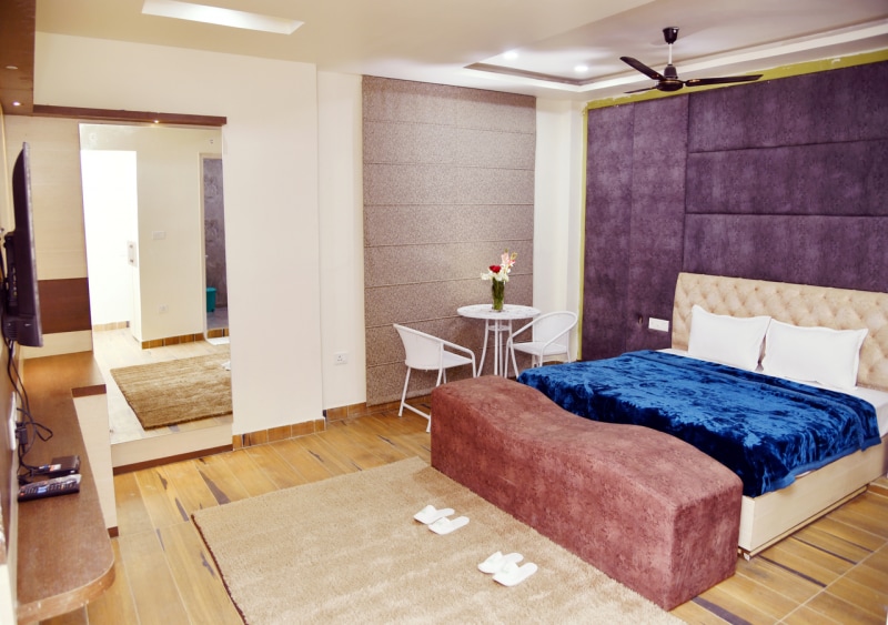 A stylish room features a spacious tufted bed covered in a blue blanket, complemented by white pillows. A cozy seating area includes a circular table and chairs, while soft rugs add warmth to the wood floor. A large mirror reflects the contemporary design.