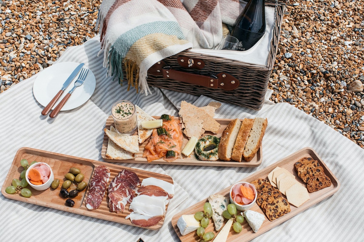 A picnic spread is arranged on a striped blanket, featuring wooden boards filled with assorted cheeses, cured meats, and fresh fruits. A wicker picnic basket sits beside the meal, accompanied by simple white plates and utensils, creating a welcoming coastal dining experience.