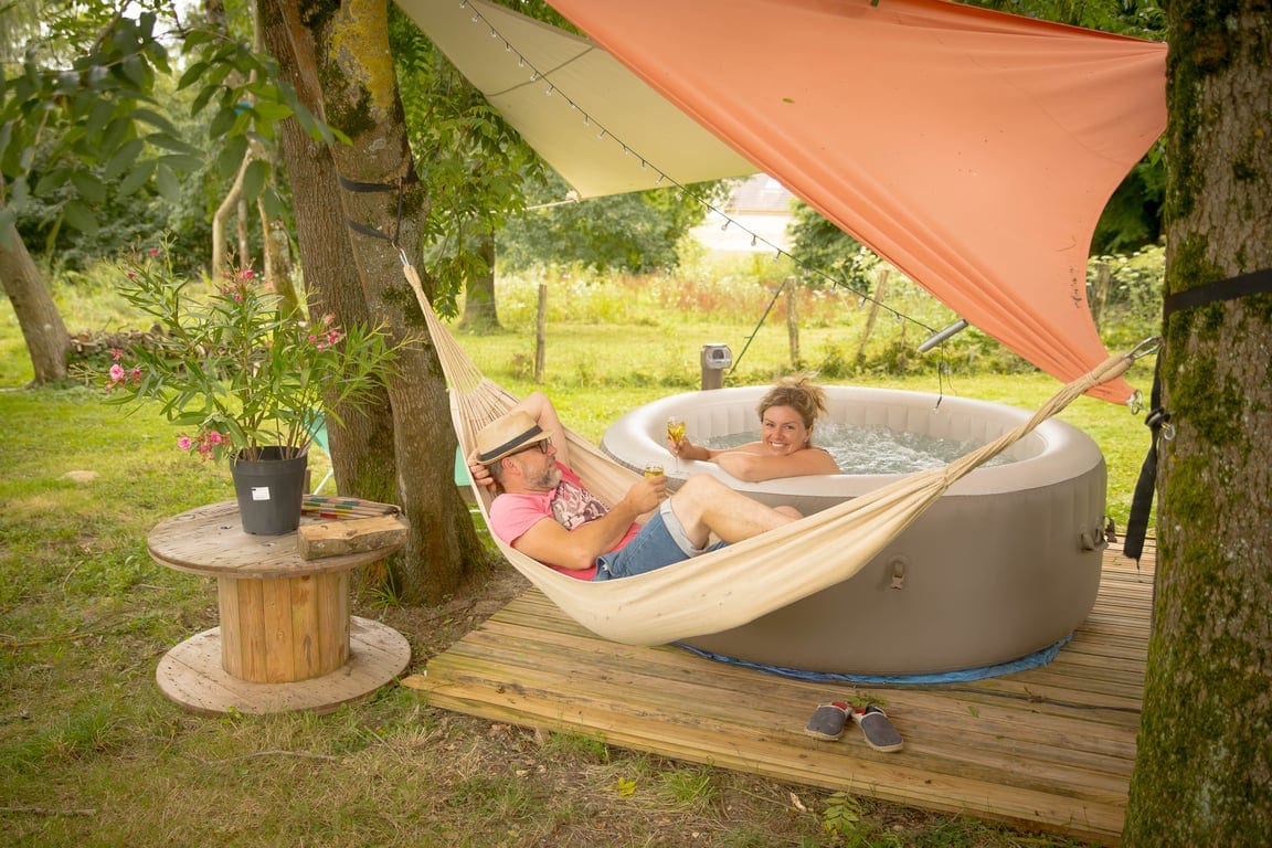 A relaxing outdoor scene features a hammock suspended between trees, with two people enjoying a hot tub nearby. A wooden table stands beside the hammock, adorned with a potted plant. The area is surrounded by lush greenery, evoking a peaceful atmosphere.