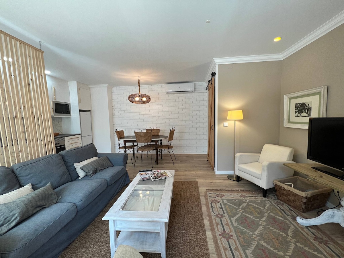 A cozy living area is showcased with a comfortable sofa and a light-colored coffee table. A dining set for four is visible in the background, alongside a well-equipped kitchen. Soft lighting enhances the inviting feel of the space.