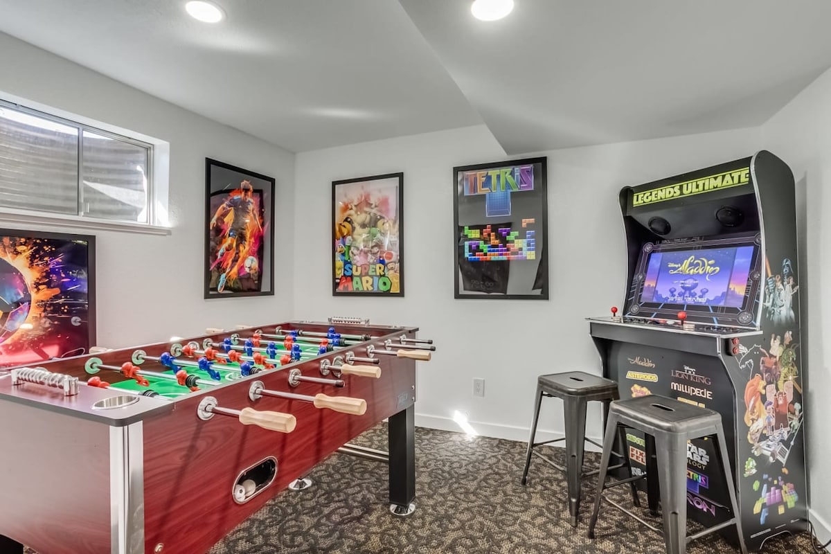 A gameroom features a foosball table and an arcade game machine displaying a selection of games. Colorful posters adorn the walls, adding a lively touch. Two barstools are positioned near the arcade cabinet, inviting guests to enjoy gaming fun.