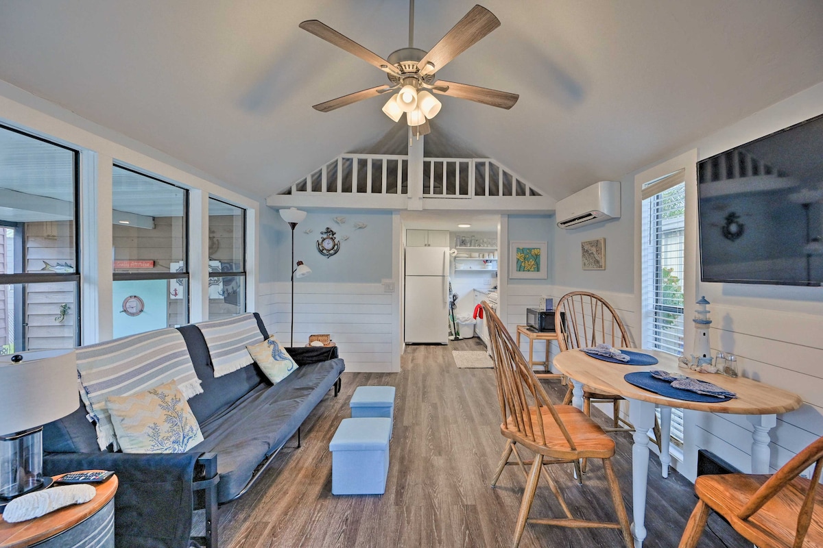 An open living space features a comfortable futon and a dining area with wooden chairs. Large windows provide natural light, and a ceiling fan enhances airflow. A kitchen area is visible in the background, showcasing modern appliances and organized countertops.