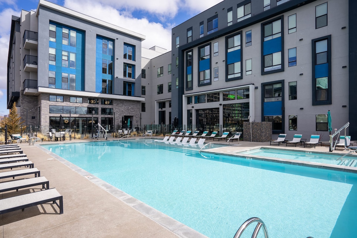 A spacious outdoor pool area features a large pool surrounded by a sun deck with lounge chairs. Modern apartment buildings in shades of blue and gray provide a contemporary backdrop, while large windows reflect natural light and views of the sky.