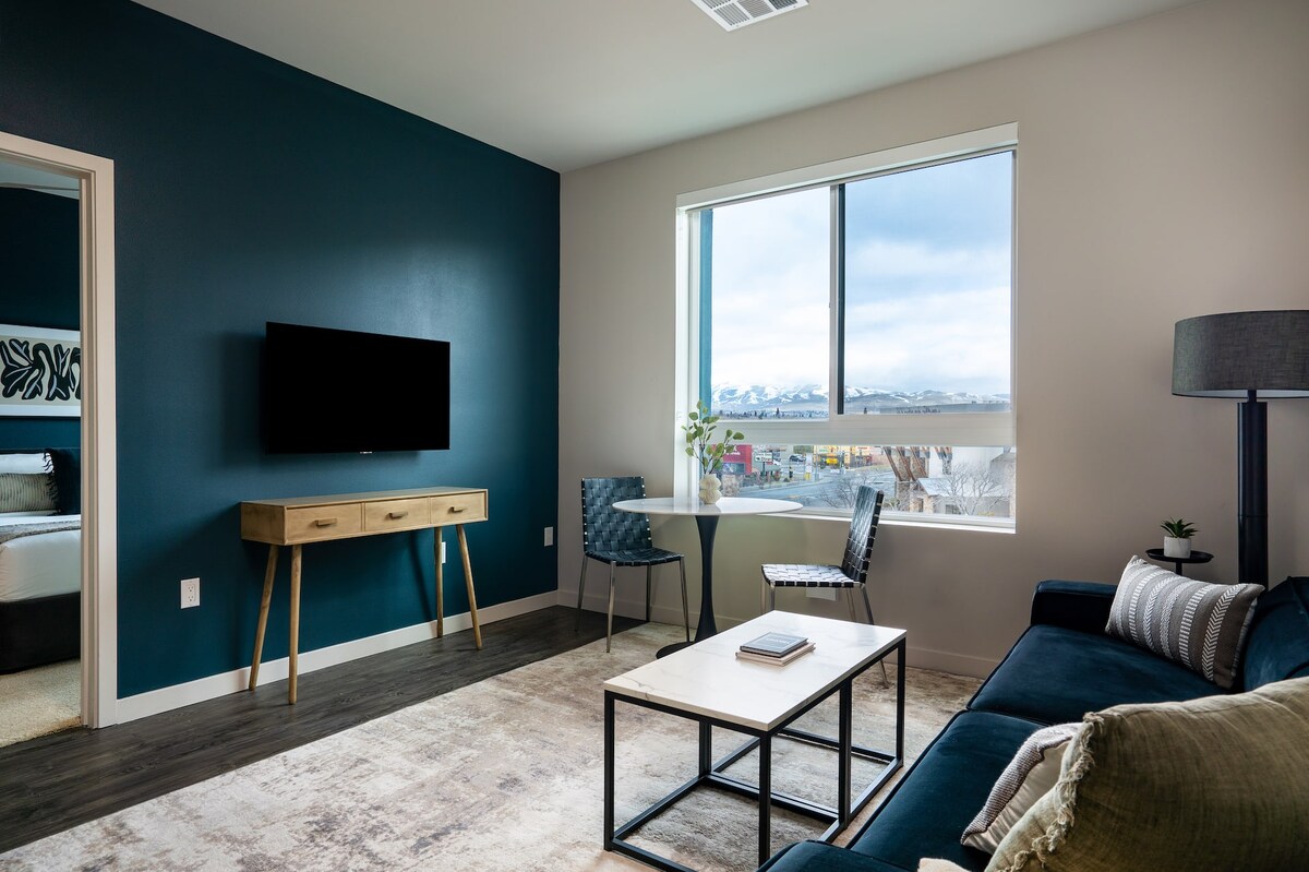 A contemporary living space is highlighted by a dark accent wall and large window offering views of the outside. A smart TV is mounted on the wall above a wooden console table. A small dining table with two chairs sits beside a cozy sofa, creating a functional and inviting area.