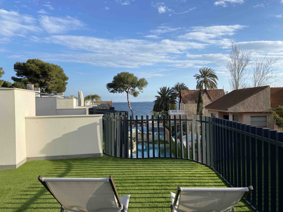 A sunlit terrace features two lounge chairs positioned on green grass, offering views of the ocean and surrounding palm trees. The horizon is visible above the fence, with glimpses of a swimming pool and nearby homes, creating a serene outdoor space.