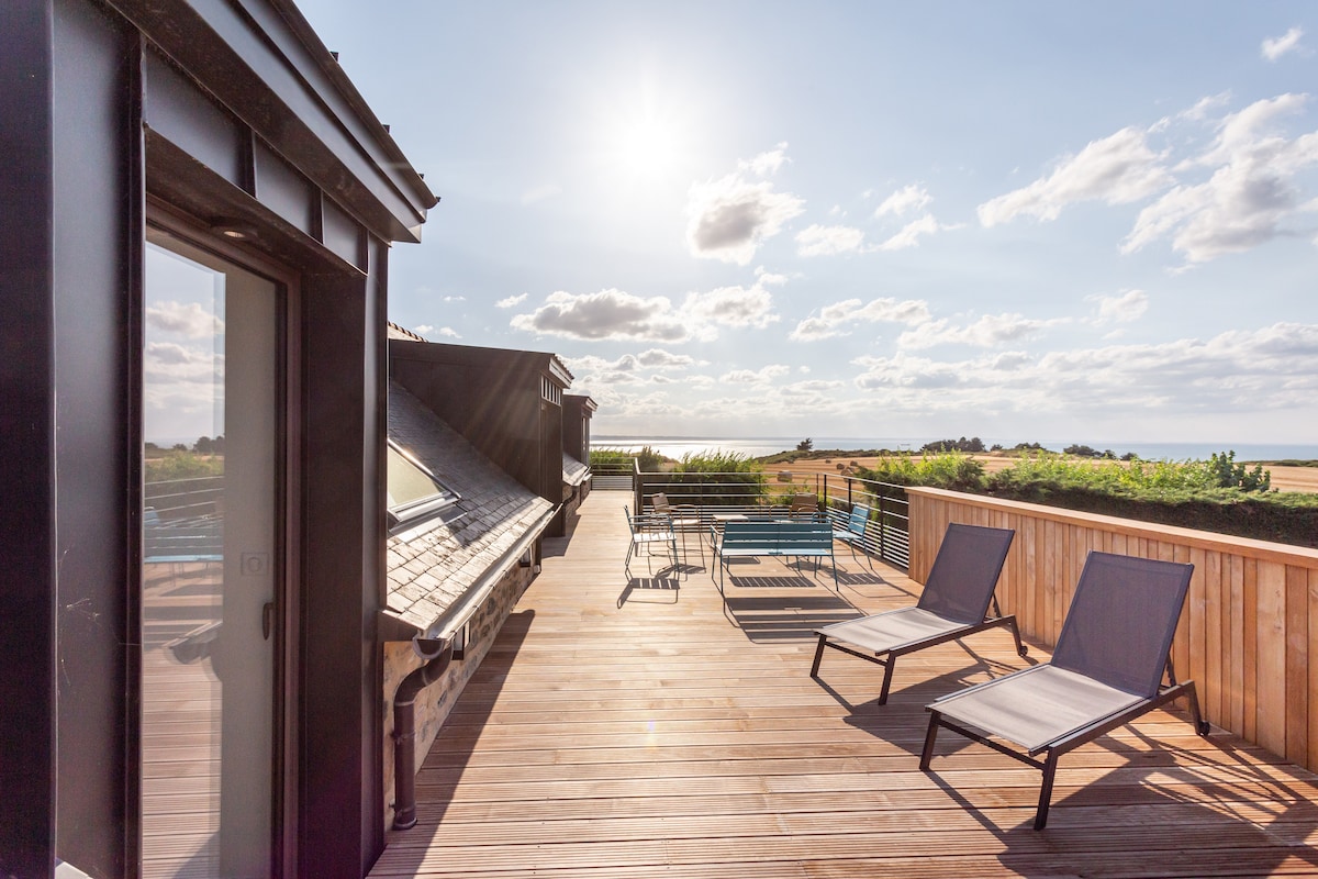 A spacious terrace is showcased, featuring two sun loungers positioned to enjoy the sunlight. The deck is surrounded by greenery, with a panoramic view of the landscape and a glimpse of the sea in the distance, inviting relaxation and outdoor enjoyment.