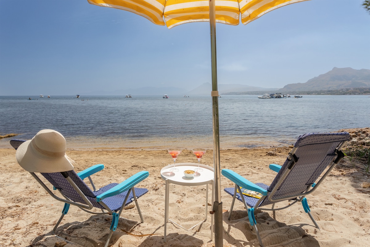 Two lounge chairs, under a striped umbrella, are positioned on sandy beachfront. A small table holds drinks, while the tranquil sea is visible in the background, dotted with boats. The sun casts a warm light over the scene, enhancing the beach atmosphere.