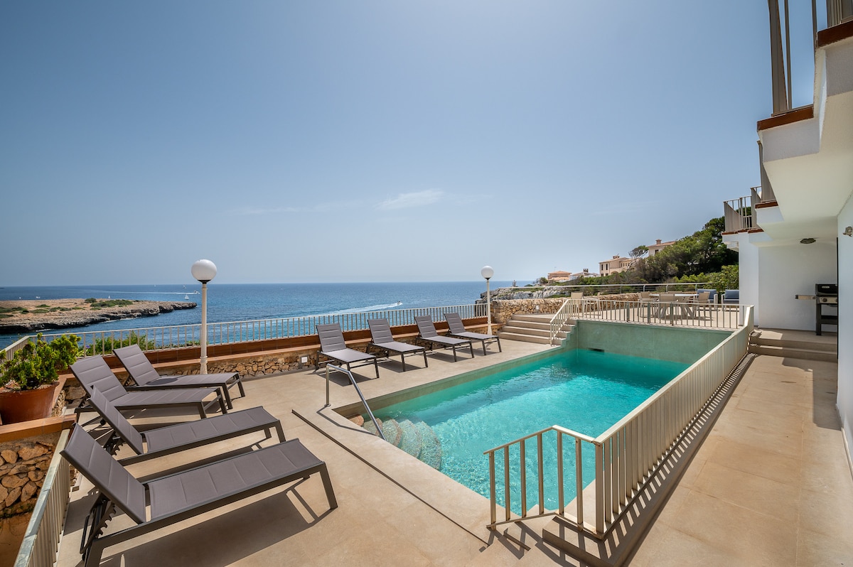 The image features a large swimming pool with clear turquoise water, set against a backdrop of the sea. Sun loungers are arranged along the poolside, and the area is equipped with protective railings. Bright sunlight enhances the serene ambiance of the outdoor space.