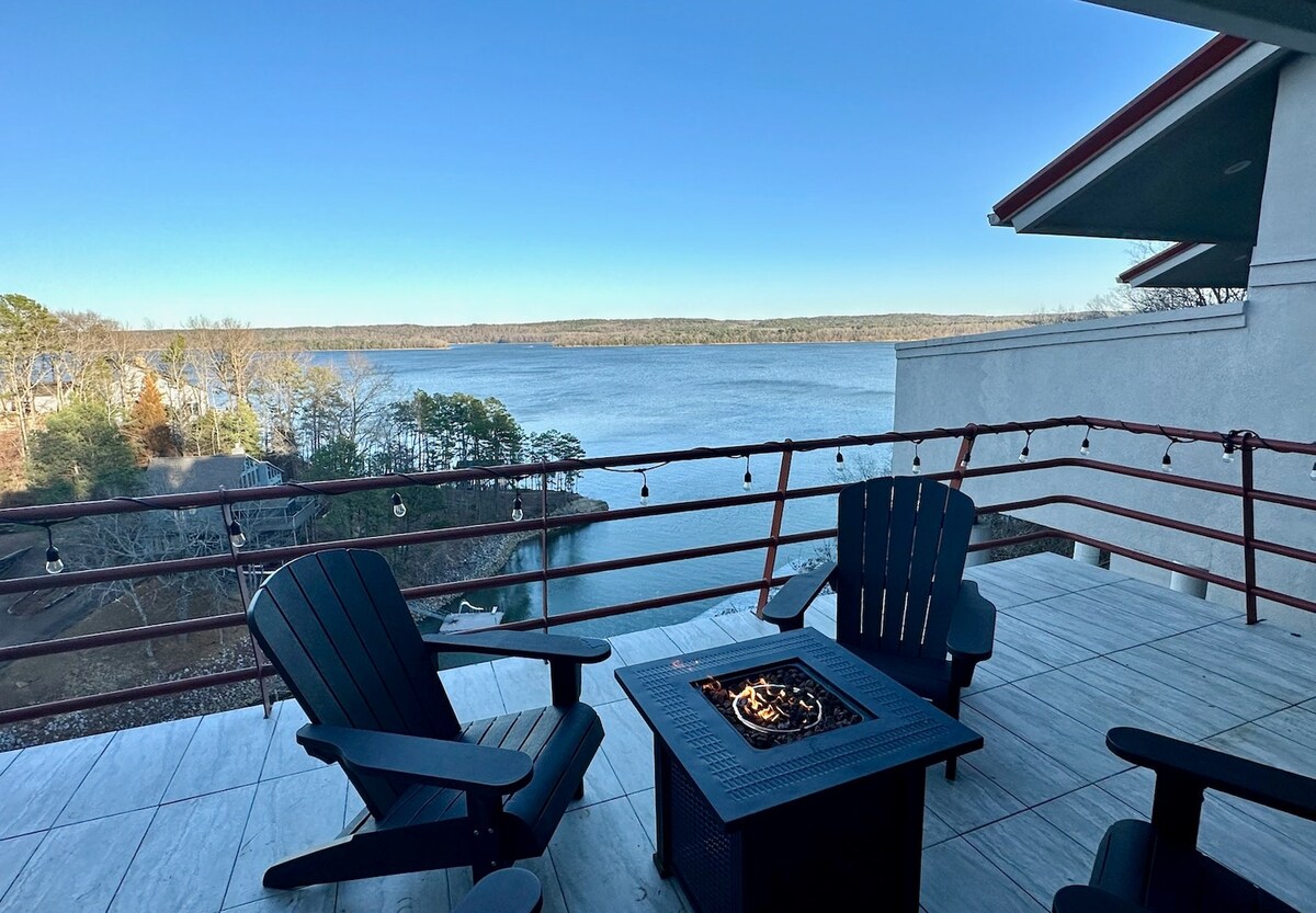 An outdoor balcony features dark wooden chairs arranged around a central fire pit. The serene lake view stretches across the horizon, framed by trees on the opposite shore. String lights add a subtle ambiance to the space, enhancing the natural surroundings.