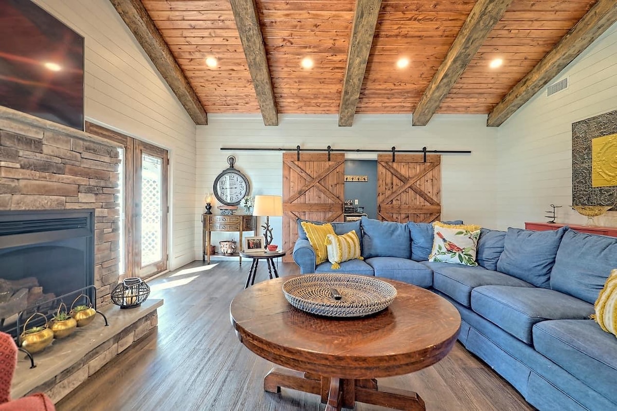 A spacious living room features a sectional blue sofa, a round wooden coffee table, and a gas fireplace framed by stone. Exposed wooden beams add warmth to the ceiling, while sliding barn doors and large windows offer an inviting touch to the room.