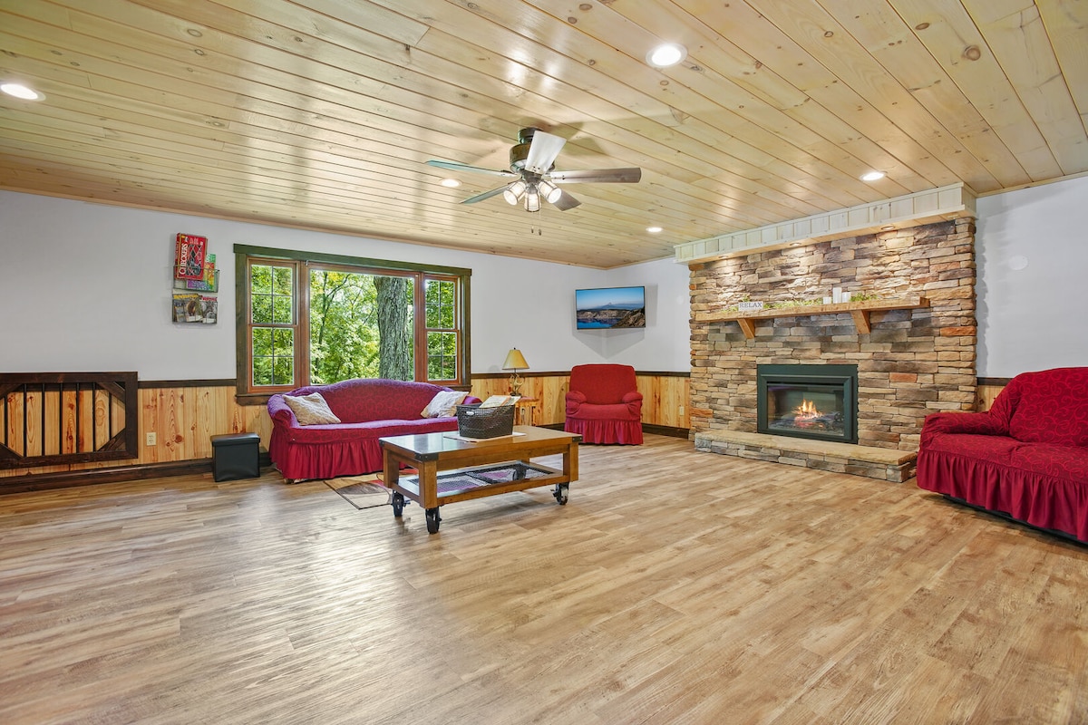 The living room features a wooden ceiling, with natural light filtering through large windows. Two red sofas and a love seat surround a central coffee table, while a stone fireplace adds a cozy focal point to the space.