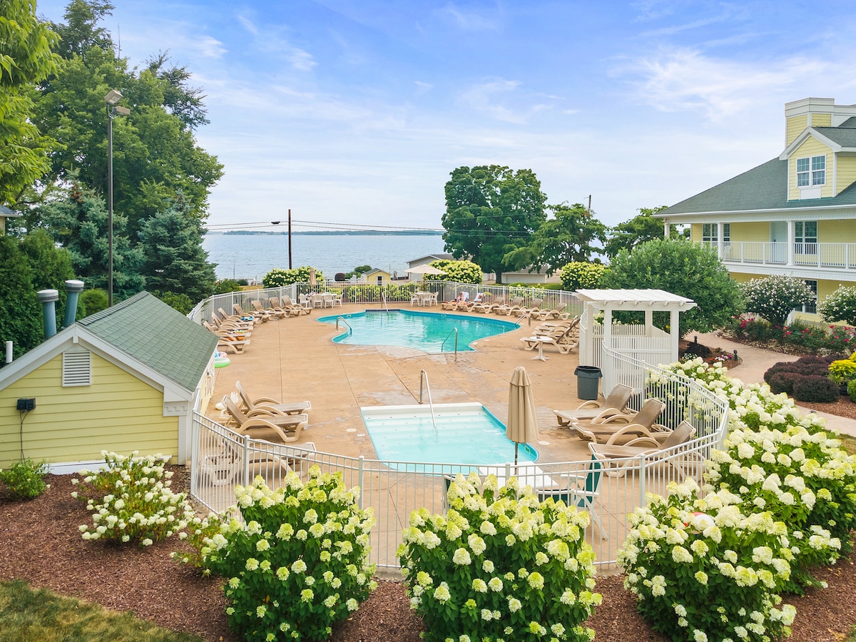 A heated outdoor pool is surrounded by a spacious deck featuring multiple lounge chairs. Lush green landscaping and blooming flowers frame the pool area. In the background, views of Lake Erie and trees enhance the serene atmosphere of the resort.