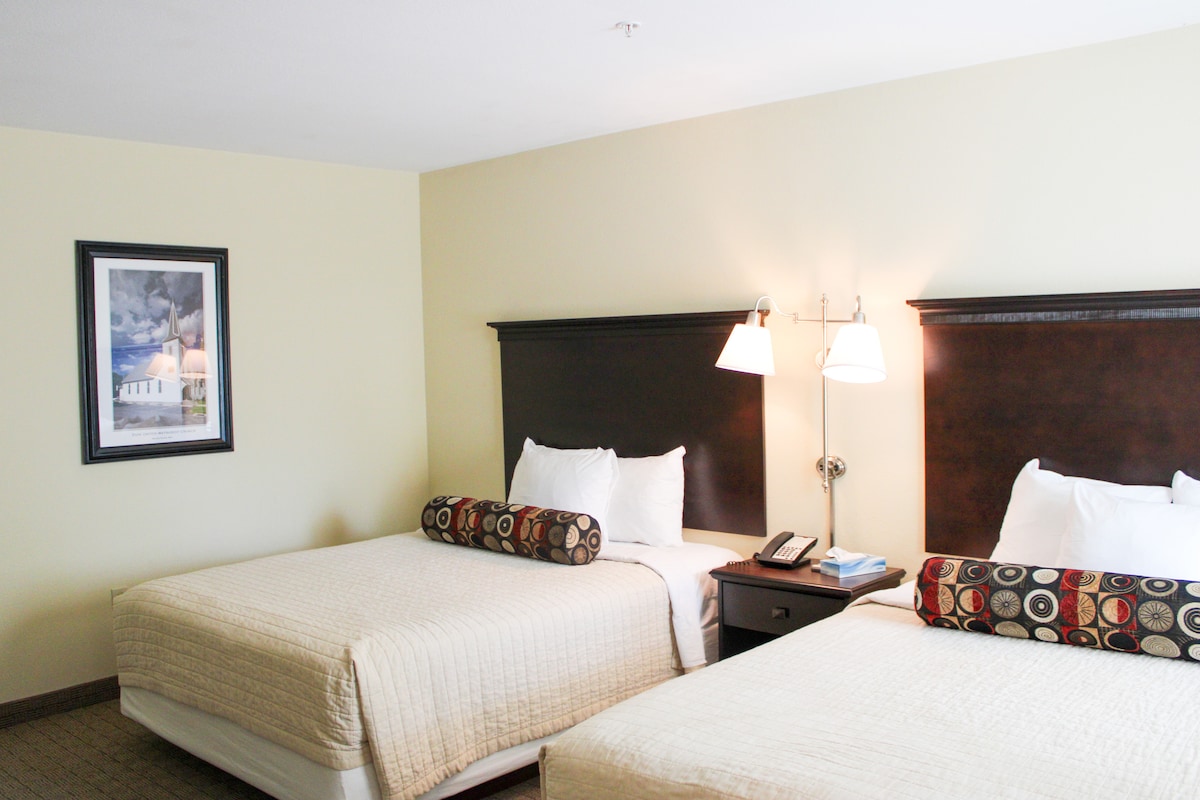 A sleeping area is shown with two queen beds, each topped with a decorative bolster pillow. Soft lighting is provided by bedside lamps mounted on the dark wooden headboards. A framed artwork piece is hung on the wall, and a nightstand sits between the beds.