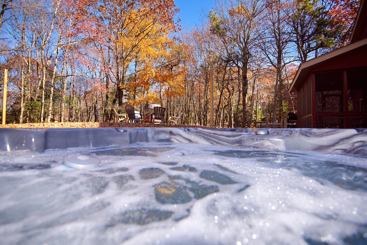 A bubbling hot tub is shown in the foreground, with a view of the colorful autumn foliage in the background. Relaxing seating is arranged in a circle nearby, surrounded by tall trees. Clear blue skies complement the serene outdoor setting.
