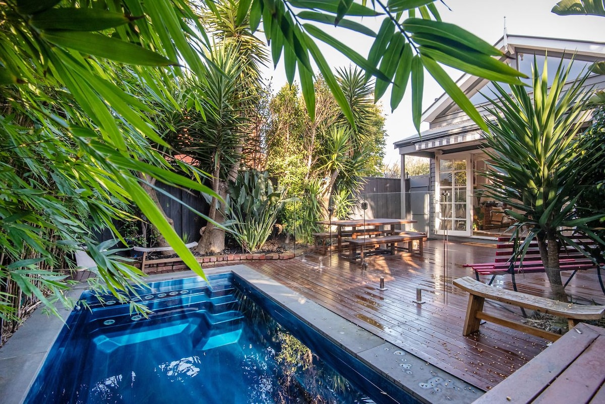 A serene outdoor space features a heated pool surrounded by tropical greenery. The wooden deck offers a dining area and additional seating. The area is designed for privacy with tall fencing and mature plants, creating a peaceful retreat.