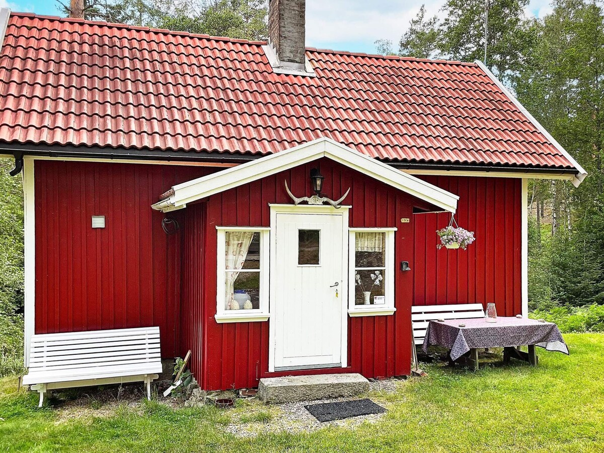 The charming red house is framed by lush greenery, featuring a white front door and two windows adorned with curtains. A small terrace with a table and chairs is visible, providing a peaceful outdoor space. Antlers decorate the entrance, adding a rustic touch.