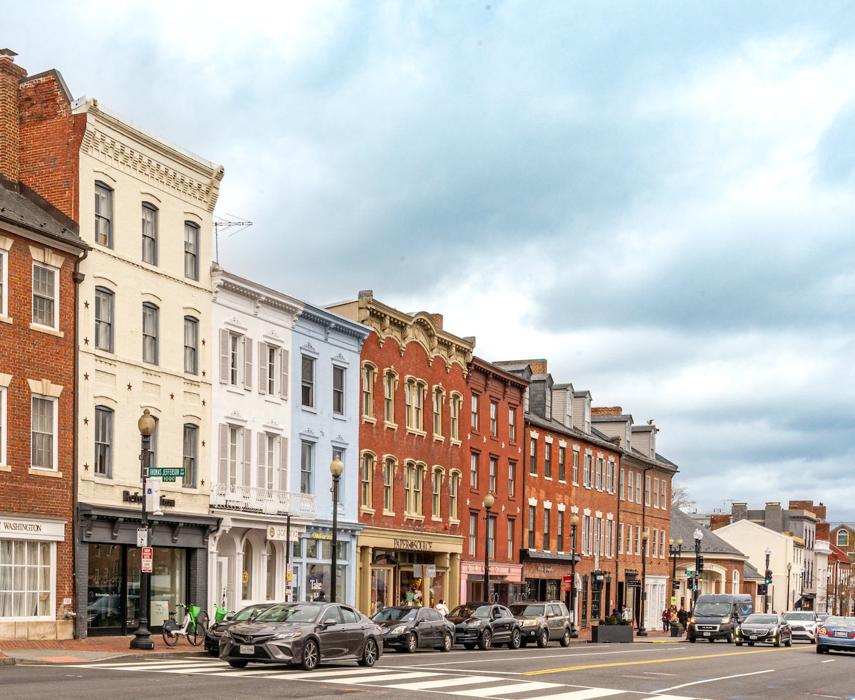 A row of historic buildings in varying hues of brick and paint lines the street. The architecture features a mix of styles with ornamental details on upper levels. Cars are parked along the street, while pedestrians can be seen walking nearby under a cloudy sky.