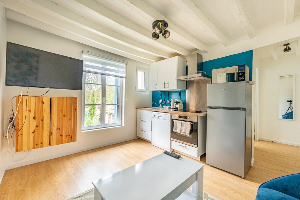A modern kitchen area includes stainless steel appliances, a refrigerator, and a microwave. A flat-screen TV is mounted on the wall, and a bright window allows natural light to fill the space. Light wood flooring complements the blue accent wall.