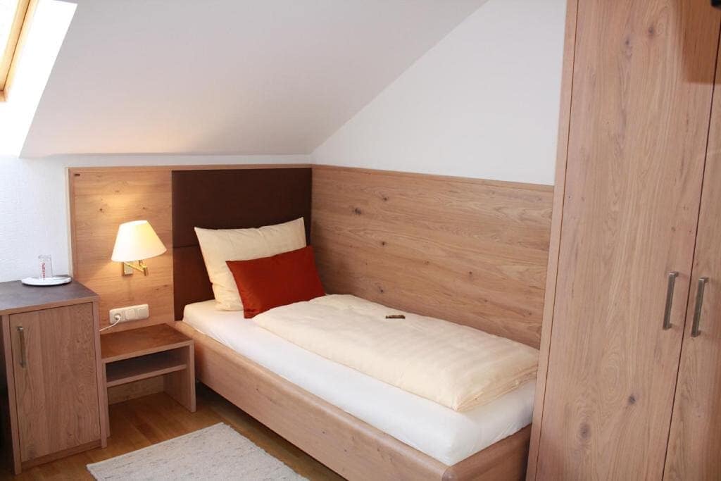 A cozy single room features a wooden bed with a neutral-colored duvet and decorative pillow. Soft light from a bedside lamp creates a warm ambiance. A small nightstand and wardrobe complement the space, while the ceiling slopes gently, enhancing the room's intimate atmosphere.