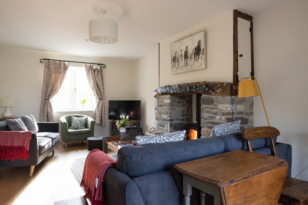 A spacious living room features a stone fireplace with a log burner. Two sofas and an armchair provide comfortable seating, while a SMART TV is positioned for entertainment. Natural light streams in through the window, complemented by soft curtains and a wooden coffee table.