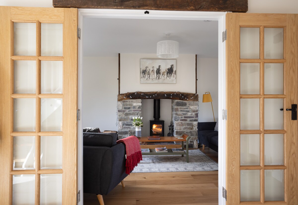 A welcoming living room is viewed through double glass doors. A stone fireplace with a log burner serves as the focal point, surrounded by two sofas and a coffee table. Light fixtures and decorative elements create a cozy ambiance, complemented by wooden beams overhead.