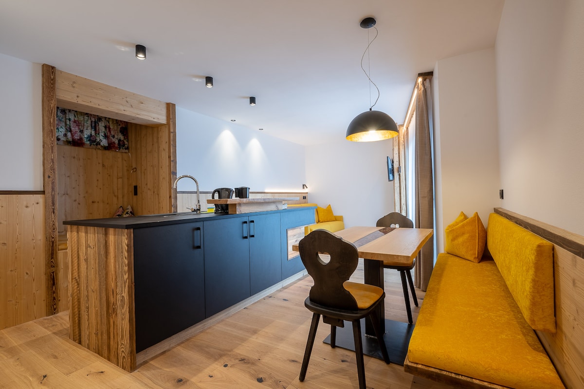 An open-plan kitchen and dining area feature a large wooden table surrounded by black chairs. A sleek black kitchen island is illuminated by modern ceiling lights. Warm wooden accents and a bright yellow seating nook add warmth to the space, while large windows offer natural light.