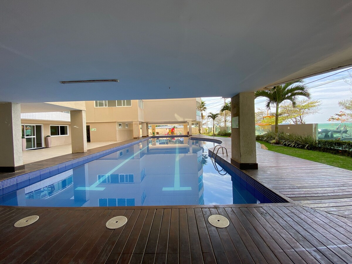A serene swimming pool is positioned under an overhang, reflecting the surrounding greenery. Sunlight filters through the open space, highlighting the blue water. Adjacent wooden decking provides an area for relaxation, while lush plants line the perimeter, enhancing the tranquil ambiance.