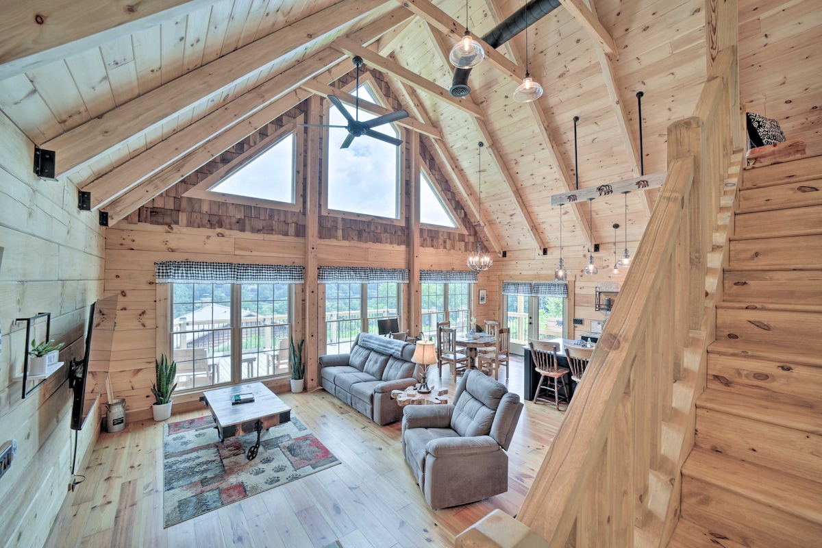 An airy great room features high wooden ceilings and large windows that reveal scenic views. Comfortable reclining chairs are positioned around a central coffee table. Light wooden walls and a cozy area rug contribute to the rustic charm, complemented by a dining area in the background.