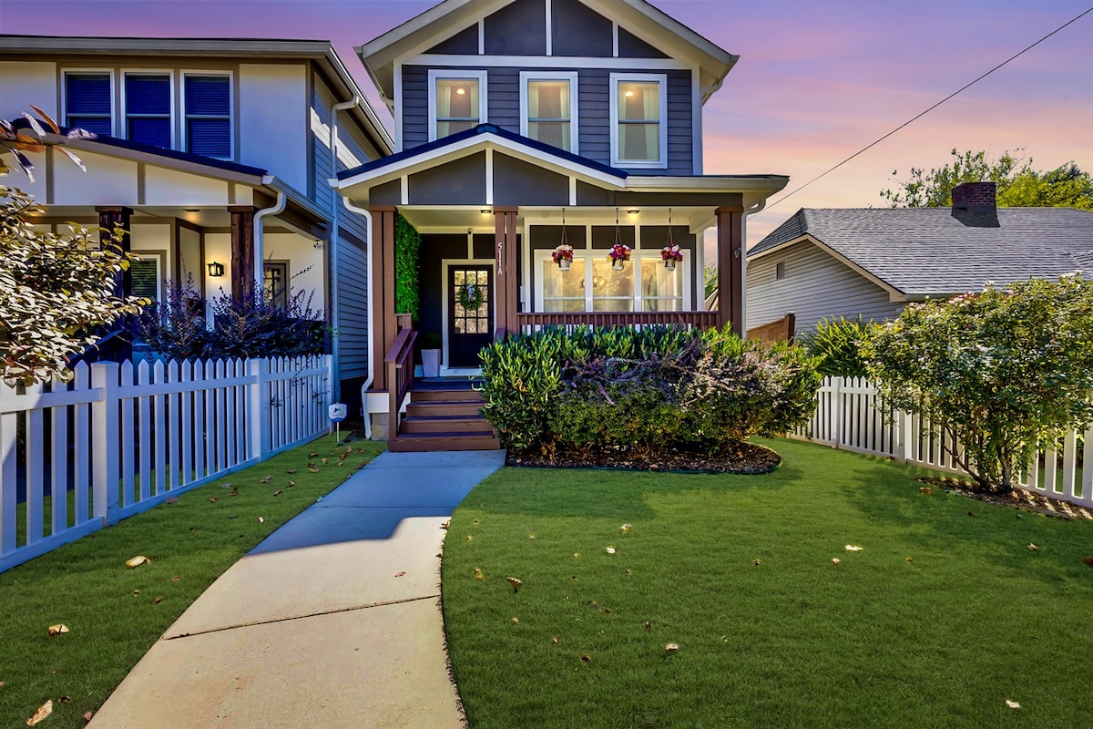 A welcoming exterior view of the home features a well-manicured front yard with green grass and a white picket fence. The entrance is highlighted by a covered porch with hanging flower baskets and a welcoming front door, framed by large windows that provide natural light.
