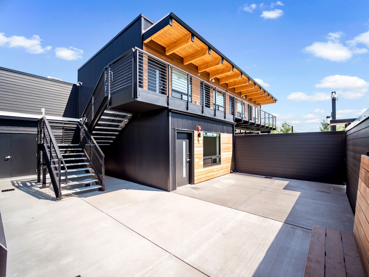The exterior of the studio unit is shown, featuring a modern design with a black facade and wooden accents. A set of stairs leads to the upper level, while an outdoor space is visible, providing concrete flooring and access to the entrance.