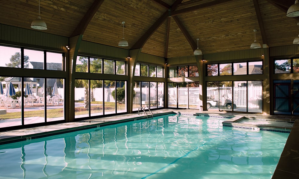 An indoor swimming pool is surrounded by large windows, allowing natural light to fill the space. The pool features a smooth, tiled perimeter and is complemented by a wooden ceiling with exposed beams. Lounge chairs can be seen outside through the glass.
