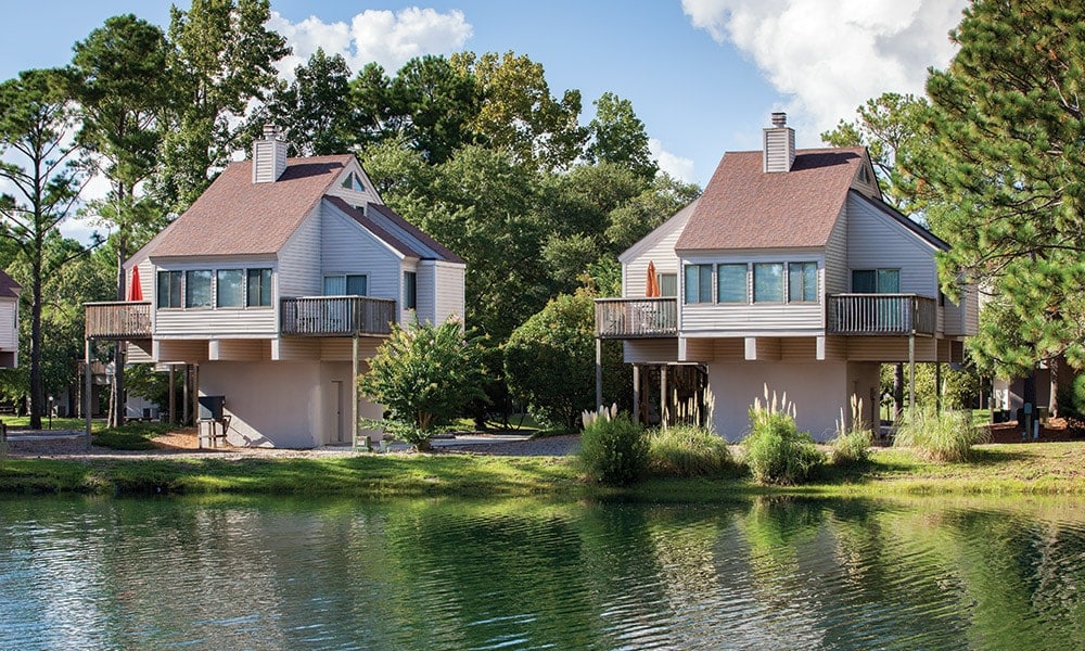 Two elevated houses are nestled beside a serene pond, surrounded by lush greenery. Each house features large windows and a deck with chairs, offering picturesque views of the outdoors. A calm atmosphere is created by the reflection of the trees on the water's surface.