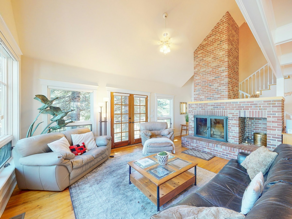 A spacious living area features high ceilings and hardwood floors, highlighted by a large brick fireplace. Natural light pours in through expansive windows, offering views of the surrounding trees. Comfortable seating arrangements include leather sofas and a wooden coffee table, creating an inviting atmosphere for relaxation.