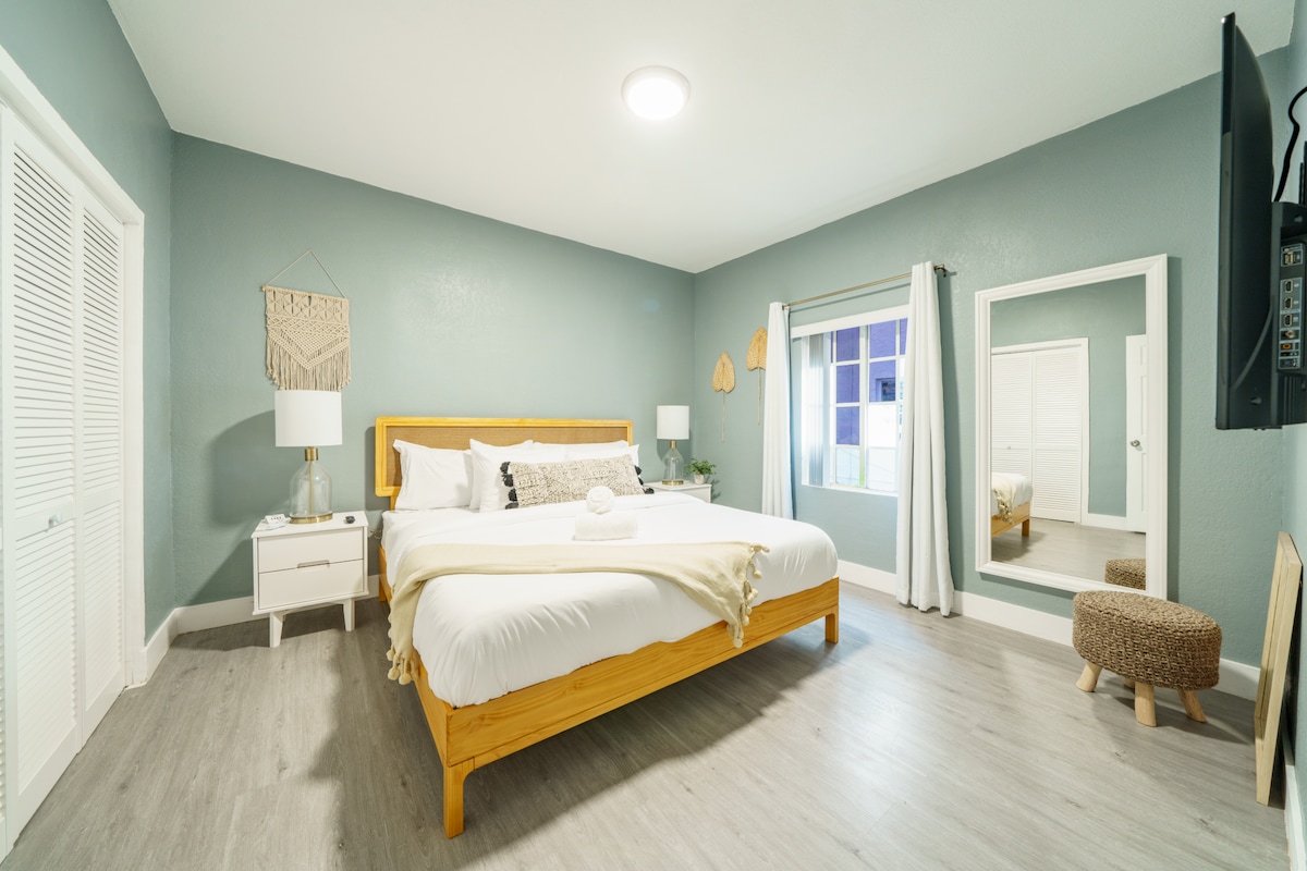 A bright and spacious bedroom features a wooden bed with soft bedding, complemented by a side table and a lamp. A large mirror stands opposite the bed, with light filtering through a window covered by sheer curtains. Light grey walls provide a serene backdrop.
