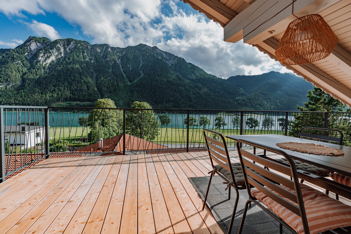 A spacious terrace is shown, featuring a dining table with several chairs. A mountain view is visible in the background, framed by clouds and greenery. The wooden deck adds a natural element, inviting outdoor relaxation near the lake.