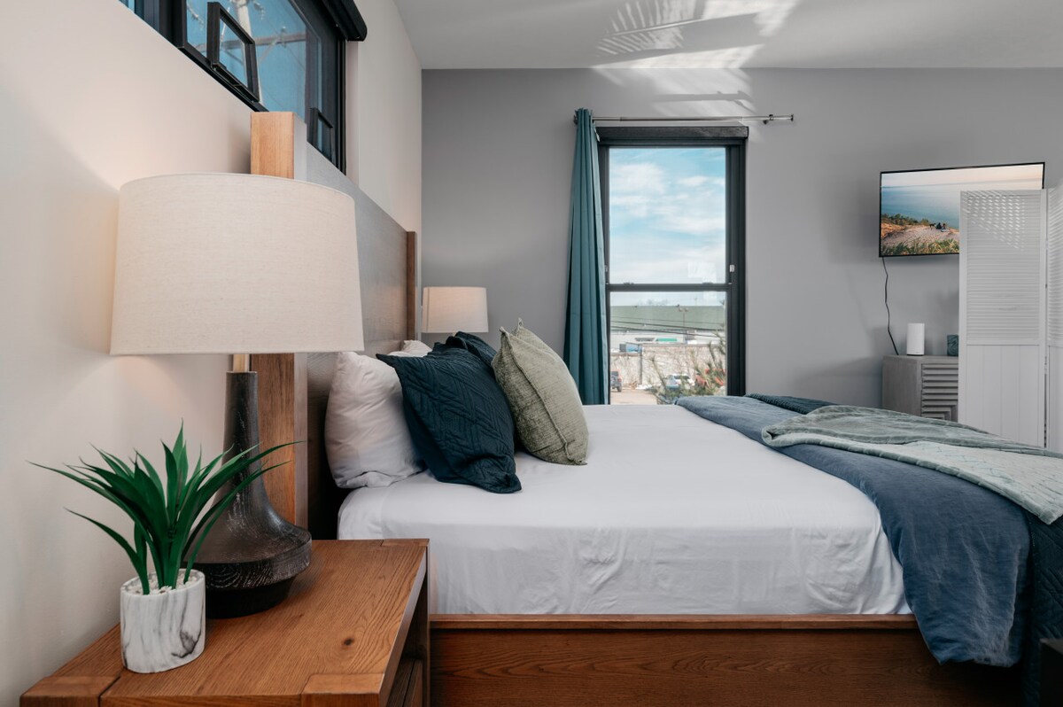 A cozy bedroom features a spacious bed adorned with dark teal and gray linens. A bedside table holds a modern lamp, and a small plant adds a touch of greenery. A large window allows natural light, with views of the outdoors visible beyond.