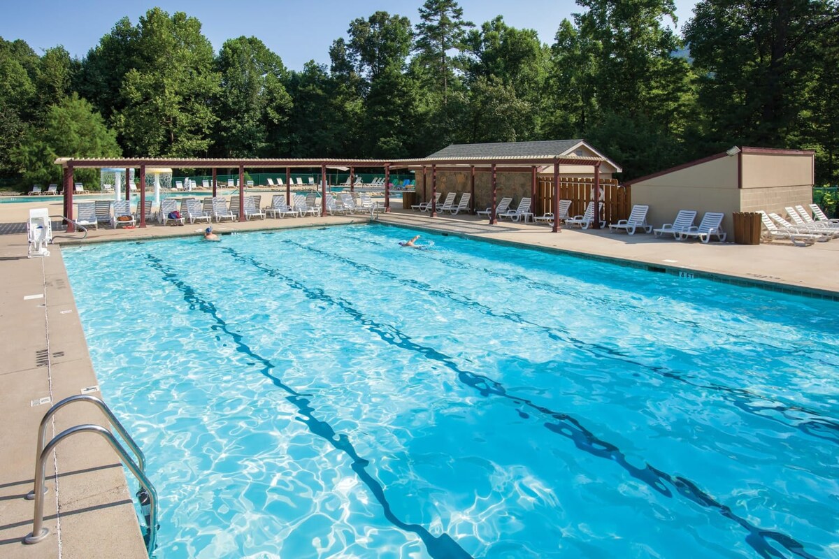A large outdoor swimming pool is visible, featuring clear blue water and lined with white lounge chairs. A shaded area and a building for restrooms or changing rooms are present nearby. Lush green trees surround the pool, creating a natural backdrop.