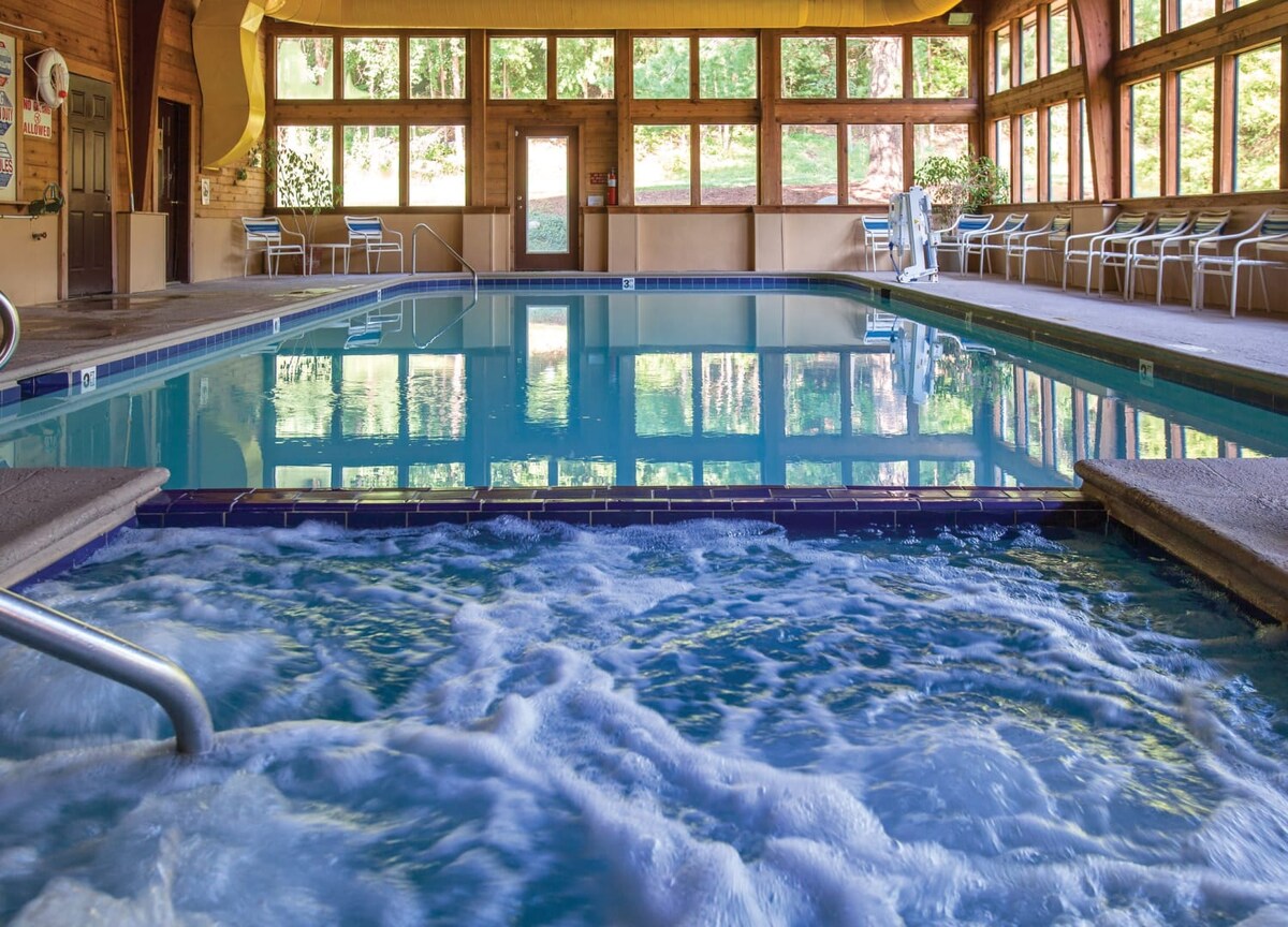An indoor swimming pool is visible, featuring clear water and a whirlpool area. Numerous windows allow natural light to fill the space, showcasing lush greenery outside. A few lounge chairs are positioned around the pool, providing a relaxing environment.