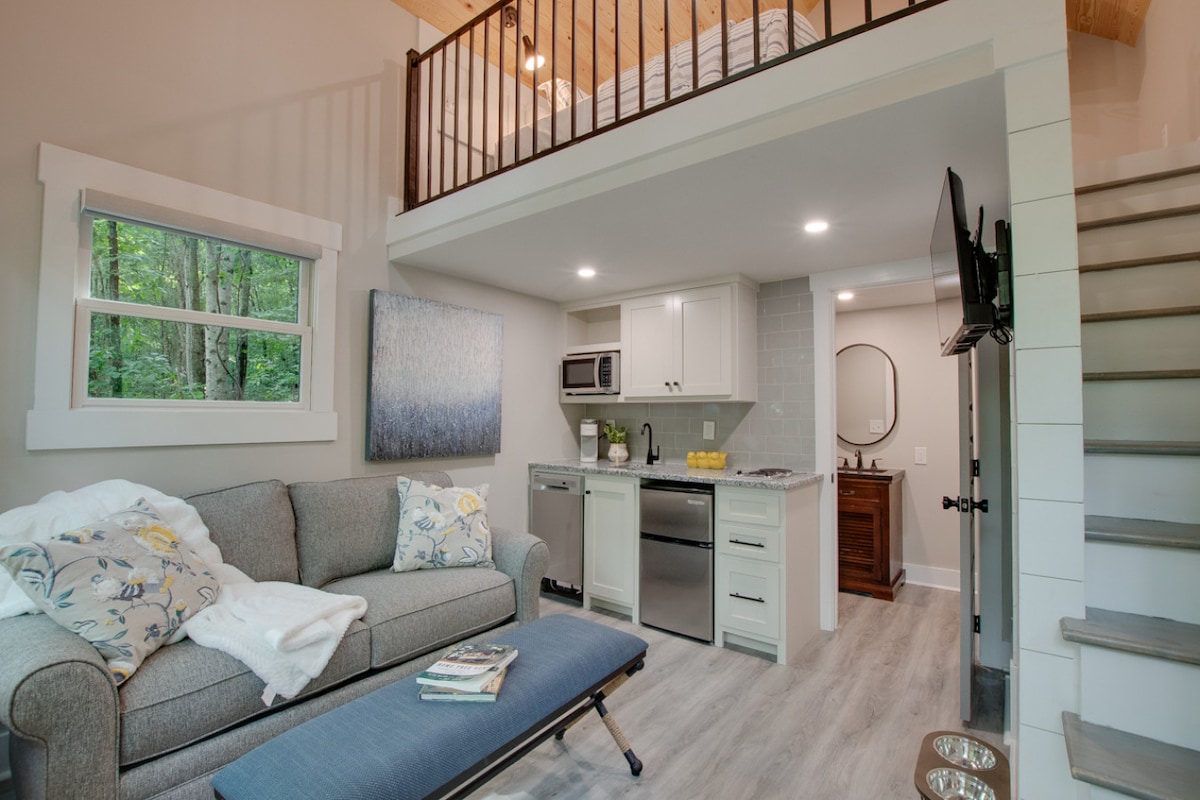 The interior features an inviting living space with a sofa and a plush bench. A kitchenette is visible, equipped with modern appliances, and a staircase leads to a lofted sleeping area. Natural light floods in through two windows, enhancing the ambiance.