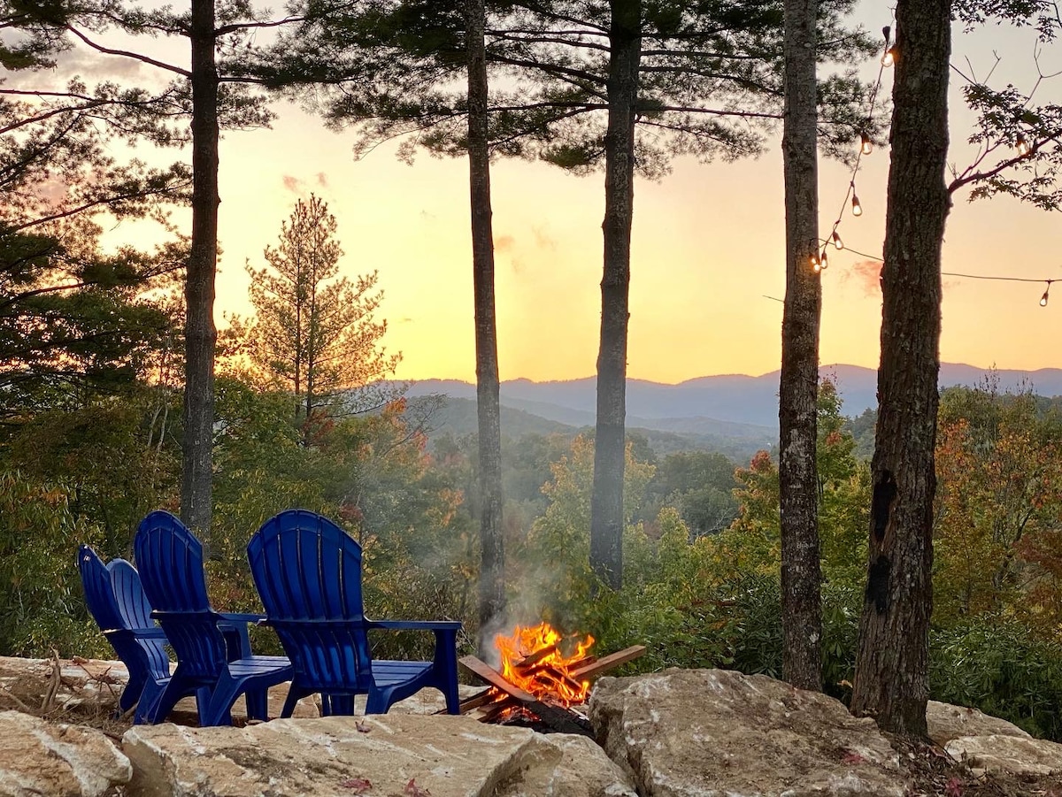 Two blue Adirondack chairs are positioned near a fire pit, surrounded by natural stone. A sunset paints the sky orange and yellow, with silhouettes of mountains in the background. Tall trees provide a sense of seclusion in this tranquil outdoor setting.