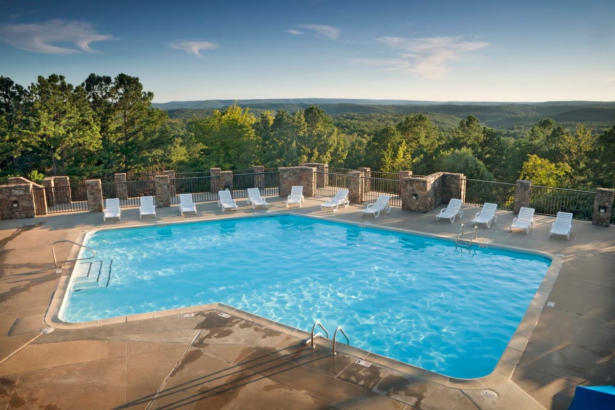 An outdoor seasonal pool is surrounded by scenic views of rolling hills and greenery. Multiple lounge chairs are positioned along the poolside, and stairs provide access to the clear blue water. The area is enclosed by a stone railing, enhancing the serene atmosphere.